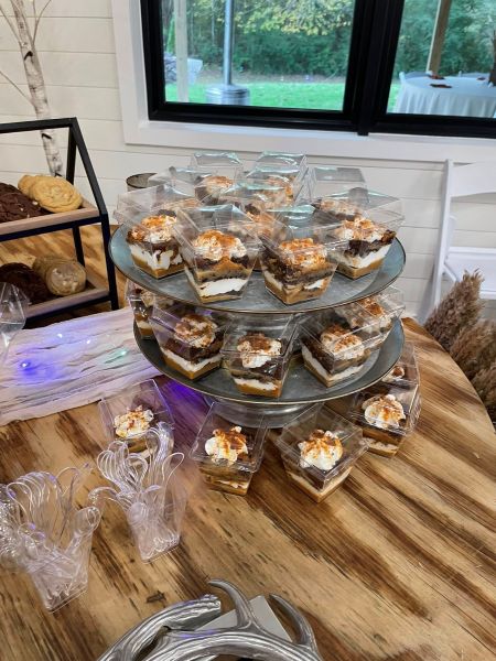 A two-tiered galvanized metal display stand showcasing individual dessert shooters. Each clear square container holds layered treats topped with whipped cream and a sprinkle of cinnamon or brown sugar. The display is set up on a rustic wooden table, accompanied by a small selection of cookies on a tiered tray and a stack of clear plastic spoons for guests to grab and go at a catered event.