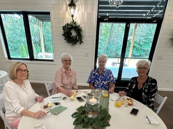 Four women seated at a round, white-clothed table in a modern farmhouse event hall, enjoying a catered brunch with elegant decor, including shiplap walls, large windows, and magnolia centerpieces.