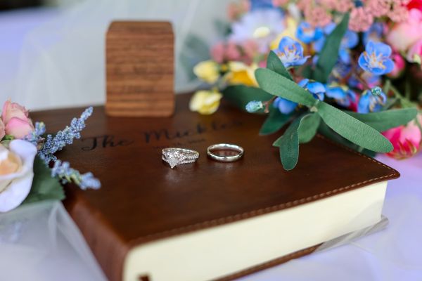 A professional close-up shot featuring a bride’s diamond engagement ring set and a groom’s wedding band resting on a personalized leather-bound guest book. The book is embossed with "The Mickles" and their wedding date. The scene is accented by a vibrant floral bouquet and a small wooden keepsake box, capturing the intimate details of a wedding day setup before the ceremony begins. This image highlights our attention to detail in styling personalized wedding mementos.