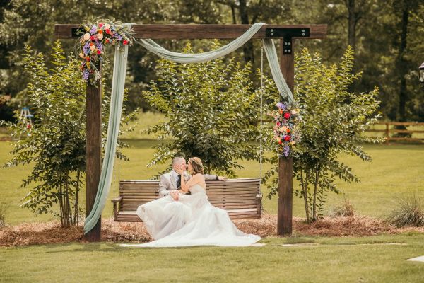 A newlywed couple shares an intimate moment sitting on our signature outdoor garden swing. The wooden structure is elegantly draped in soft sage fabric and adorned with vibrant, multicolored floral arrangements, perfectly framing the bride and groom. This picturesque spot, nestled within our lush green venue grounds, provides a romantic and timeless backdrop ideal for wedding portraits that capture the essence of a serene, nature-inspired ceremony.