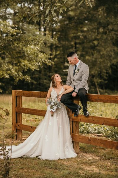 A newlywed couple sharing an intimate, loving moment on our property. The bride, in a detailed lace gown, looks up at her groom, who is perched on our signature wooden fence. The natural woodland backdrop and rustic fence highlight the outdoor beauty of our venue, capturing the timeless and relaxed atmosphere we offer for wedding portraits.