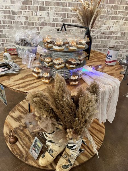 A tiered display featuring individual clear cups layered with chocolate cake, cream, and caramel, topped with a dusting of cocoa. The desserts are arranged on a rustic wooden spool table decorated with dried pampas grass, vintage-style metal decor, and soft fairy lights. This setup highlights our signature "Rustic Dessert Station" aesthetic, perfect for farm-themed weddings or barn events.