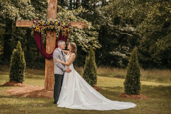 A bride and groom standing before a rustic wooden cross decorated with burgundy draping and floral accents at an outdoor wedding ceremony.