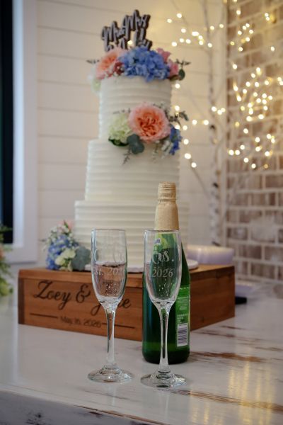 A centerpiece shot featuring a three-tiered white wedding cake decorated with fresh pastel flowers and a "Mr. & Mrs." cake topper. In the foreground, two engraved champagne flutes marked "Est. 2026" and a chilled bottle of champagne are positioned on a personalized wooden stand engraved with the couple's names and wedding date, "Zoey & [Name], May 16, 2026." The background features a rustic white brick wall illuminated by warm fairy lights, creating an elegant and celebratory atmosphere for our wedding reception.