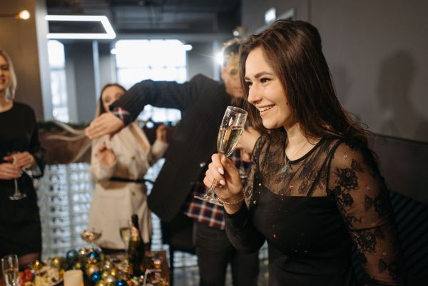 A young woman in a black lace dress holds a glass of champagne, smiling during a festive office holiday mixer at an event venue, with colleagues socializing in the background.