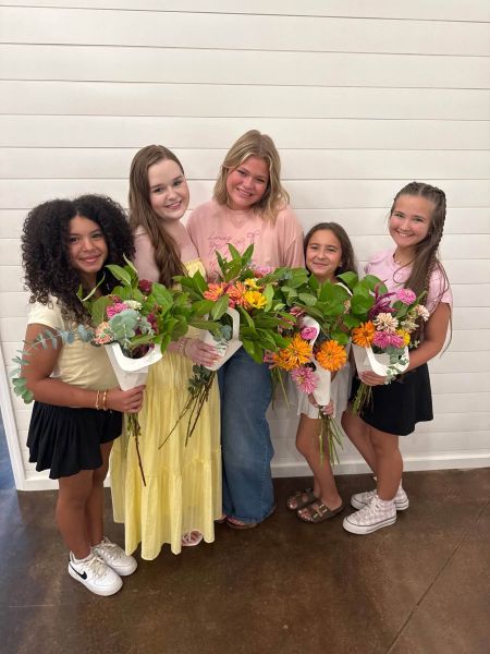Five smiling young participants standing together against a white shiplap wall, each proudly holding a unique, freshly arranged bouquet from our floral workshop. The arrangements feature bright zinnias, eucalyptus, and other seasonal greenery in white paper wraps. This photo highlights the joy of creative expression and the beautiful, custom floral designs crafted by our students during their visit to our studio.