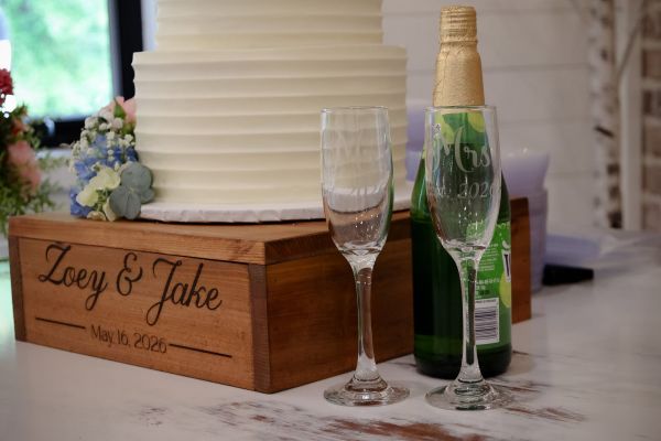 A close-up shot of a custom-engraved wooden display crate featuring the names "Zoey & Jake" and their wedding date, May 16, 2026. Atop the crate sits a tiered, minimalist white wedding cake accented with fresh floral sprays. Beside the cake are two engraved champagne flutes labeled "Mr." and "Mrs." alongside a bottle of sparkling cider, ready for the couple’s celebratory toast. This image captures the intimate, personalized details we coordinate for wedding receptions at our venue.
