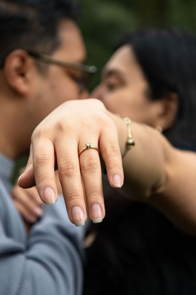 A close-up shot focusing on a delicate gold engagement ring with a central diamond stone, worn on a woman's hand. The background features a soft-focus, blurred image of a couple embracing, capturing an intimate and romantic moment. This image highlights the classic elegance of our fine jewelry in a real-life, celebratory context, perfect for showcasing our commitment to commemorating special milestones.