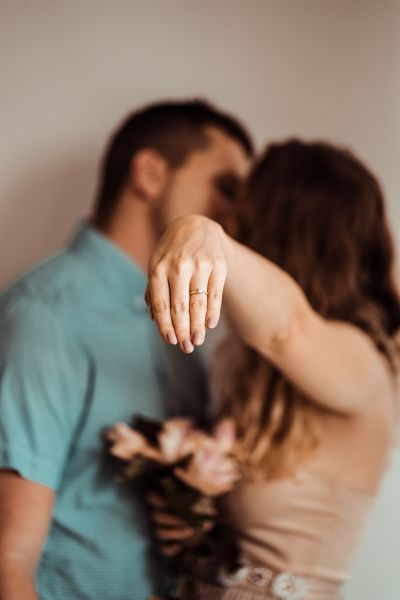A close-up focus on a delicate gold solitaire diamond engagement ring worn on a woman's hand, positioned in the foreground. The background features a softly blurred, romantic silhouette of a couple embracing, capturing the intimate emotion of a proposal. This image perfectly highlights the craftsmanship of our jewelry as the center of a life-changing moment.