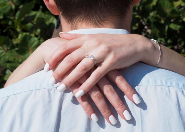 A close-up shot focusing on a woman’s hands wrapped around a partner's neck in an intimate embrace. Her left hand is prominently featured, showcasing an elegant engagement ring with a round center stone and a diamond-pave band. Her nails are manicured with a clean, solid white polish, contrasting with the soft natural lighting and green foliage in the background. This image perfectly captures the romantic sentiment of a proposal or engagement, ideal for highlighting our luxury jewelry collections.