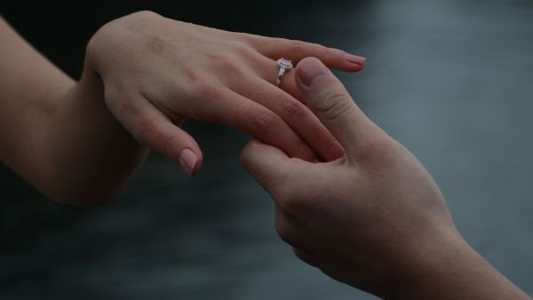 A close-up shot focusing on a delicate hand wearing a signature engagement ring, capturing the tender, intimate moment of two hands holding to emphasize the timeless nature of the jewelry. The soft, moody background highlights the brilliance of the center stone.